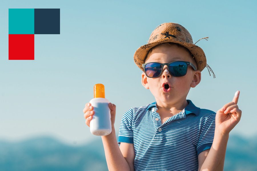 Kleiner Junge mit Hut und Sonnenbrille hat eine Sonnencreme in der Hand und einen Klecks Sonnencreme auf der Nase.